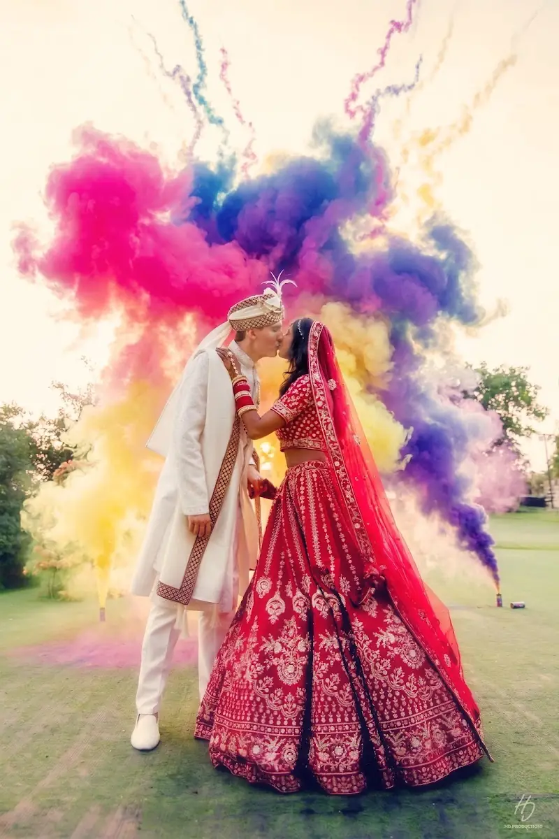 A bride in a red lehenga and a groom in a cream sherwani and turban kissing, surrounded by a burst of pink, purple, yellow and blue coloured smoke on a lawn.