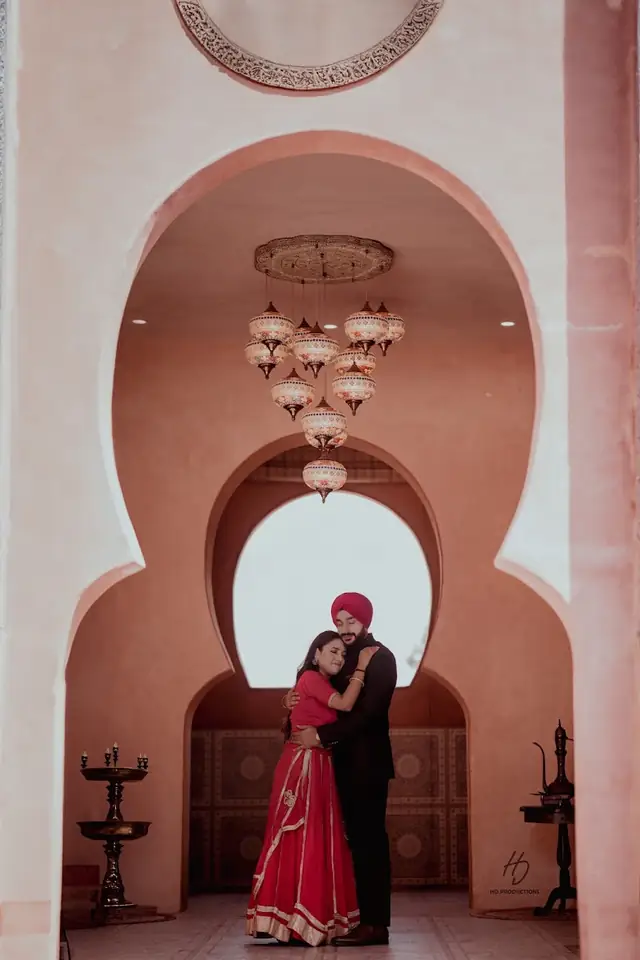 A couple embracing under a series of Moroccan-style arches hung with patterned glass lanterns, the groom in a maroon turban and black sherwani, the bride in a red and gold lehenga.