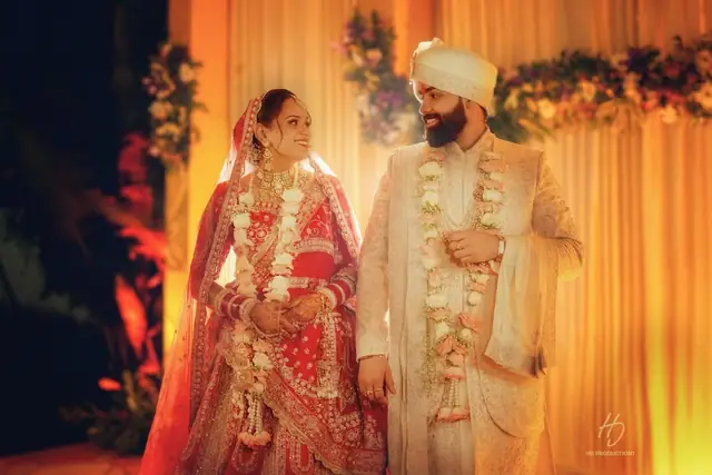A bride in a red and gold lehenga with a dupatta over her head and a groom in a cream sherwani and turban, both smiling at each other, floral garlands around their necks, in front of a marigold and rose backdrop.