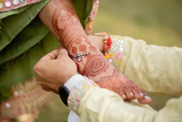 Close-up of a hennaed hand and forearm being held by another hand, a silver charm bracelet on the wrist, against a green background.