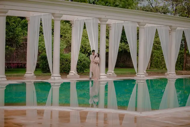 A couple standing between white colonnade pillars beside a turquoise pool, their reflection mirrored in the water.