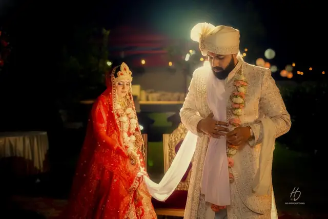 A bride in a red lehenga and gold crown-style tikka standing beside a groom in a cream sherwani and turban holding her dupatta, lit warmly at night.