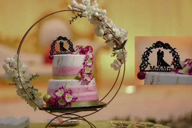 A two-tier pink and white wedding cake decorated with sugar flowers on a gold ring stand, with a silhouette 'Mr & Mrs' cake topper.