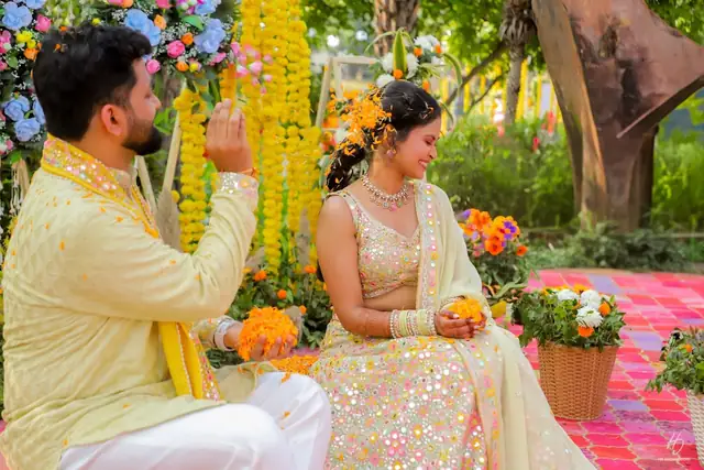 A groom in a yellow kurta showering marigold petals over a bride in a mirror-work lehenga, both seated outdoors amid hanging marigold garlands.